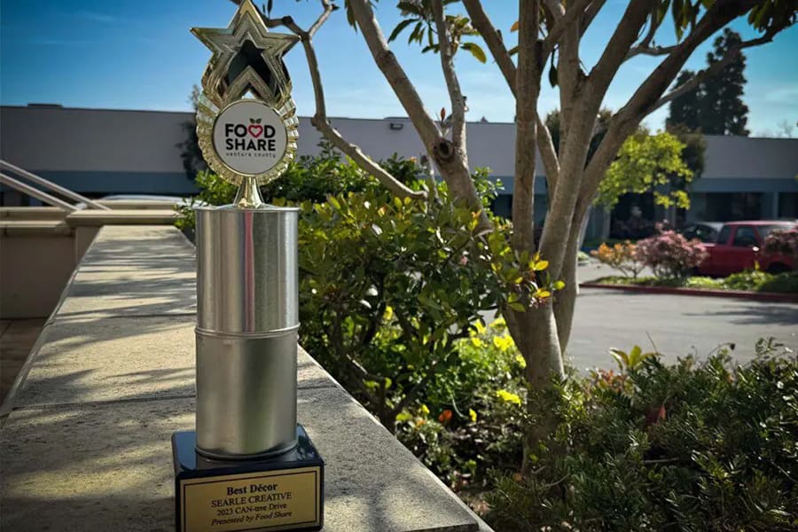 A "Best Decor" trophy awarded to Searle Creative by Food Share is displayed outdoors on a stone ledge, with trees and a building visible in the background. This marks another proud win for Searle Creative at the CAN-Tree event.