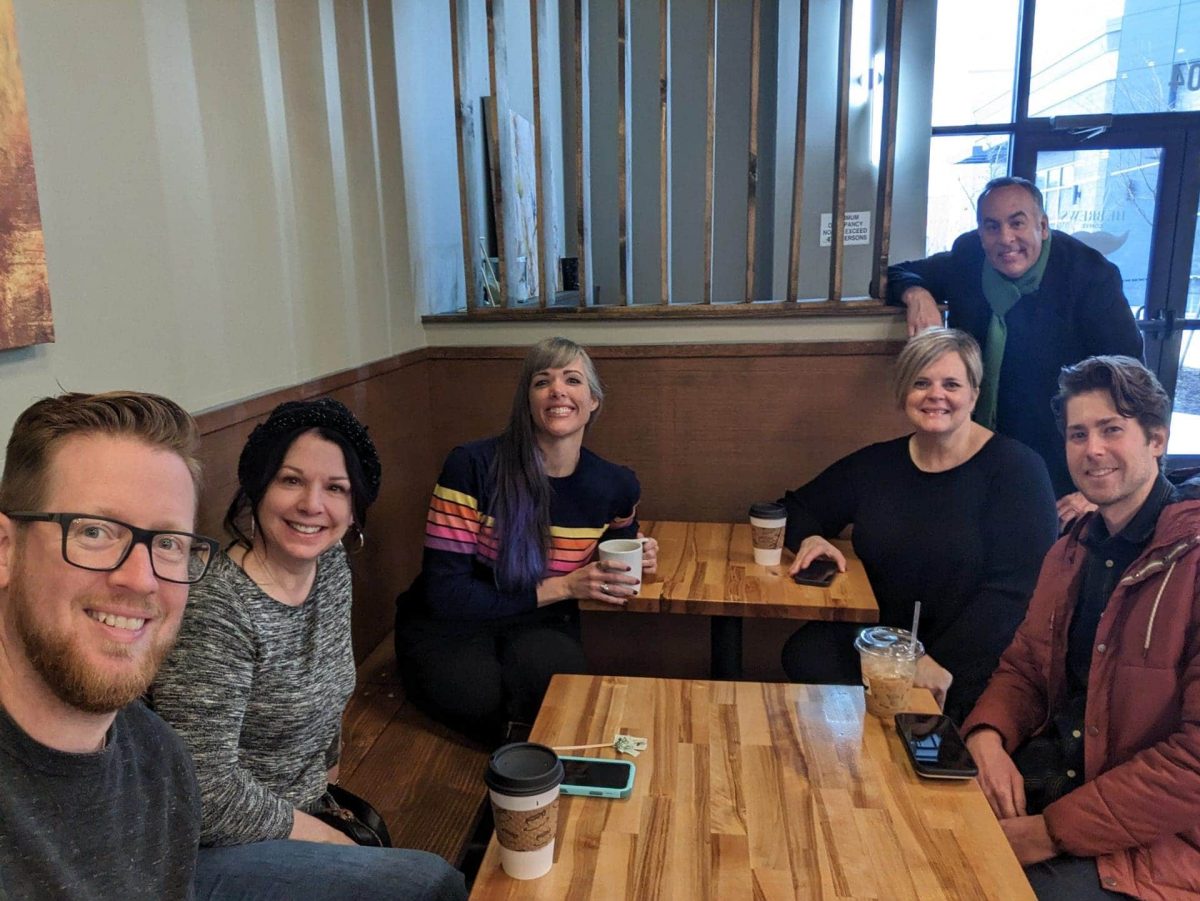 A team of six people sits at a wooden table in a cafe, smiling and holding coffee cups.