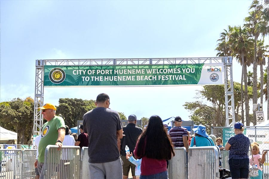 HBF23_EntranceSign People entering the Hueneme Beach Festival under a welcome banner with text "City of Port Hueneme Welcomes You to the Hueneme Beach Festival." Palm trees and a speed limit sign are visible in the background.
