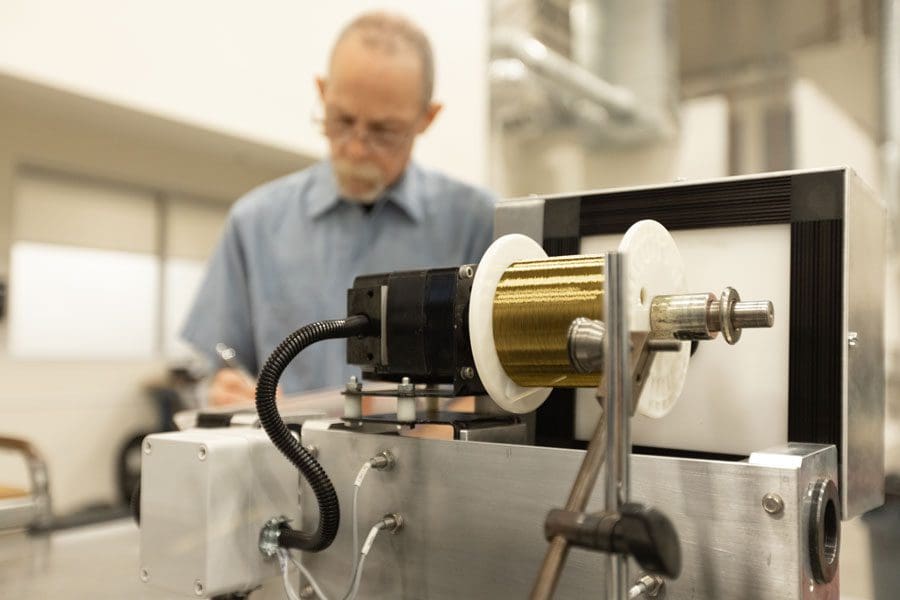 A man in a workshop is writing in the background. In the foreground, captured in the photo, is industrial equipment with a spool of golden wire. A solid demonstration of manufacturing video and photo production.