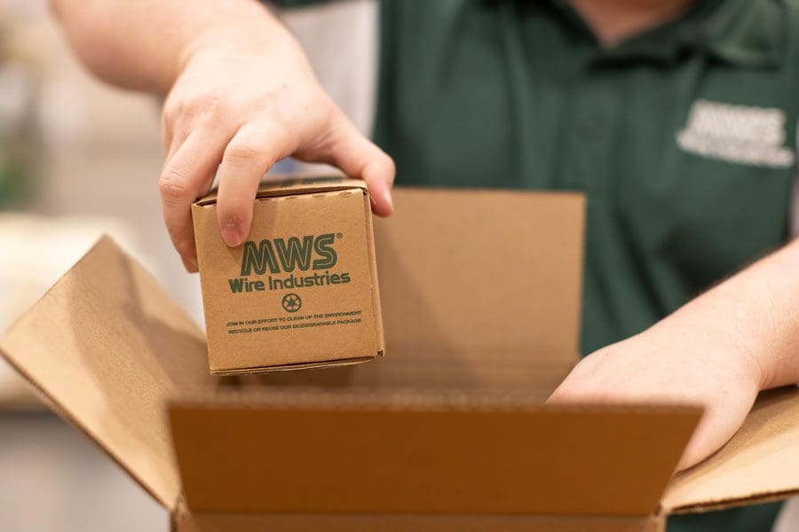 A person in a green shirt is meticulously placing a small cardboard box labeled "MWS Wire Industries" into a larger open cardboard box, staged perfectly for manufacturing video and photo production.