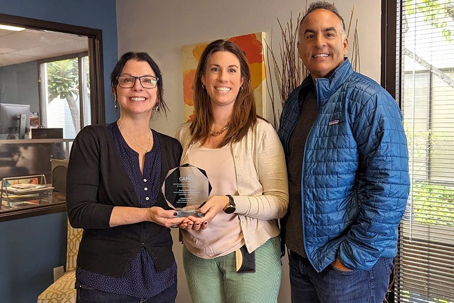 Three people stand indoors, smiling and holding a translucent CAPIO EDGE Award. They are dressed in casual business attire, in an office setting with a window and artwork in the background, representing the collaborative triumph of Searle Creative Group.