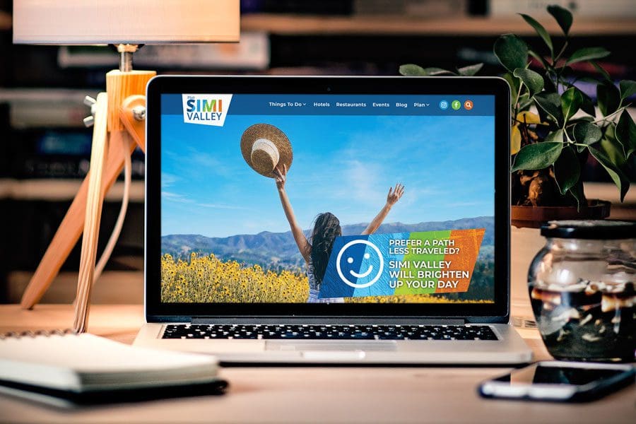 A laptop on a desk displays the Simi Valley Website, showcasing an image of a woman in a field of yellow flowers holding a hat with mountains in the background.