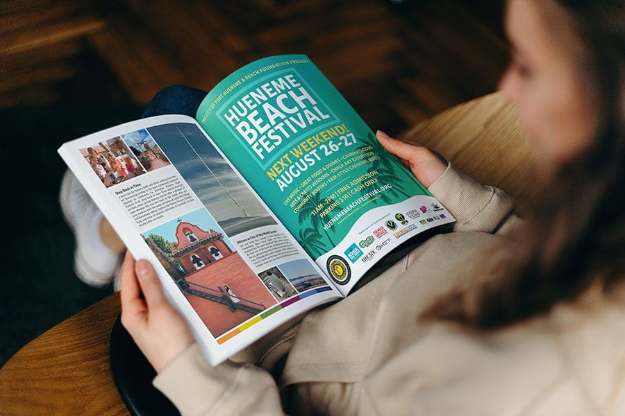 smartmockups_lphcf5cf-reduced Person reading an open magazine featuring an advertisement for the Hueneme Beach Festival happening next weekend, August 26-27. The page includes event details and colorful photos of the lively festival atmosphere.