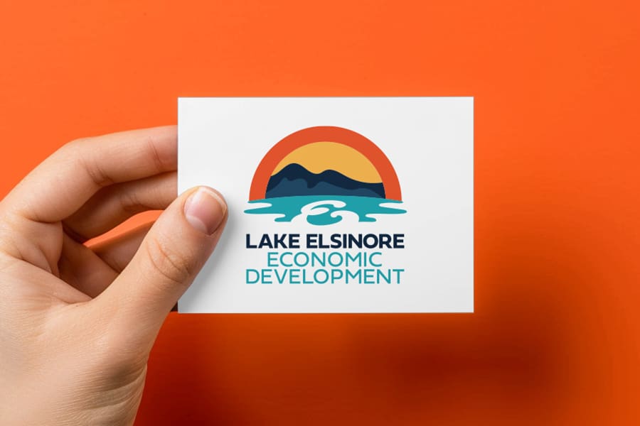 LEED_Portfolio_01 A hand holds a vibrant card displaying the Lake Elsinore Economic Development logo, featuring a sunset, majestic mountains, and a serene lake on an orange background.