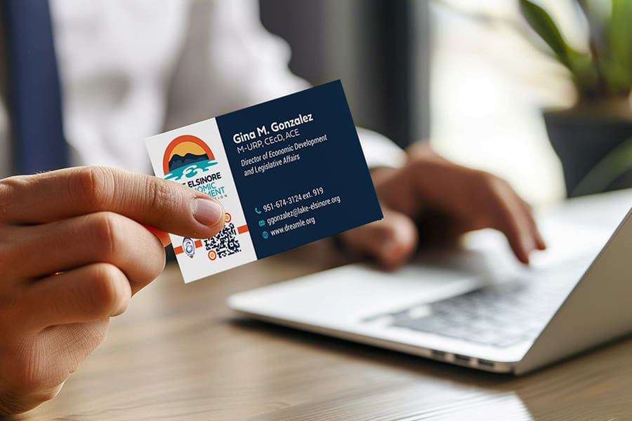 LEED_Portfolio_02 Person holding a business card showcasing a vibrant logo alongside their name, title, and contact details. In the background, a laptop displays information about economic development in Lake Elsinore.