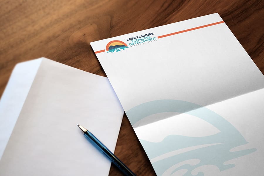 LEED_Portfolio_05 A blank letterhead featuring the Lake Elsinore Development logo sits on a wooden surface next to an unsealed envelope and a blue pen, embodying the essence of strategic branding for economic development in the heart of Lake Elsinore.