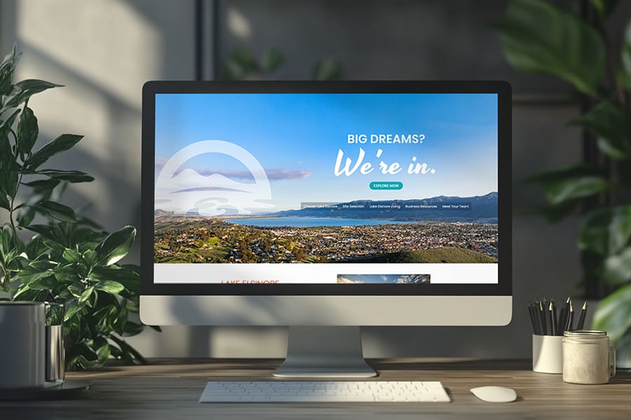 A desktop computer on a wooden desk showcases the Economic Development website, featuring a panoramic cityscape and the text BIG DREAMS? We're in. It's surrounded by indoor plants and soft natural lighting, adding a touch of Lake Elsinore charm.