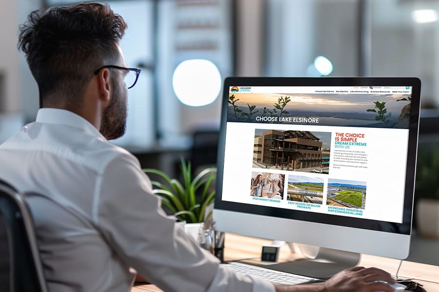 A man in a white shirt sits at his desk, exploring a website dedicated to the economic development of Lake Elsinore on his desktop computer screen.