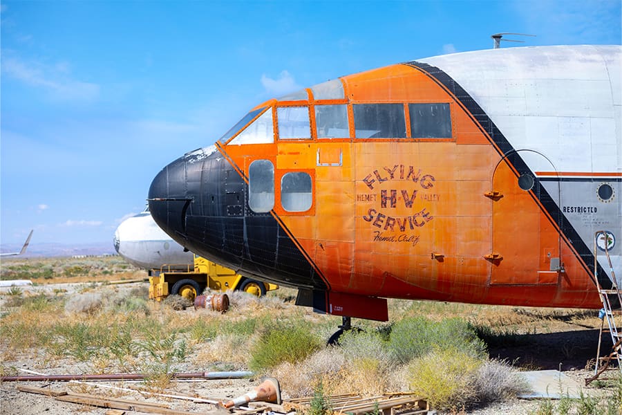 An abandoned orange and black airplane, bearing Flying HW Service on its side, rests in a dry field with scattered vegetation—a captivating subject for photography enthusiasts exploring Lancaster Economic Development opportunities.