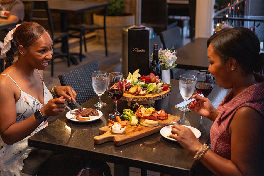 Under the warm glow of elegant lighting, two people dine at a restaurant table adorned with steak, wine, and a charcuterie board. Their lively conversation is captured like a perfect moment in photography, highlighting the vibrant atmosphere that Lancaster Economic Development proudly supports.