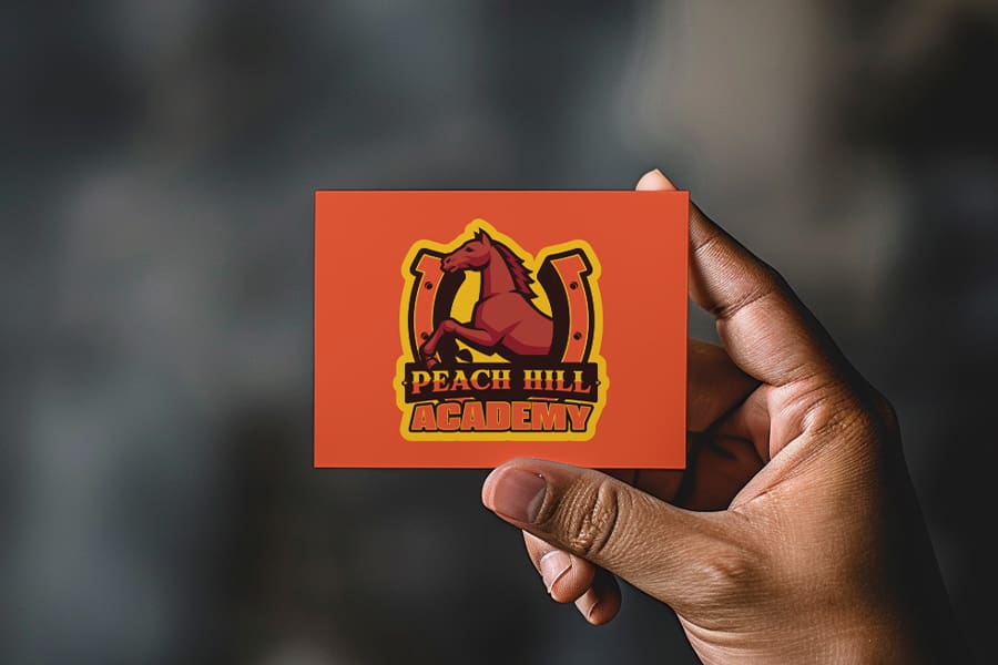 MUSD_PeachHillAcademy_Logo00 A hand holds a card showcasing the Academy Logo of Peach Hill Academy, featuring a horse and horseshoe on an orange background.