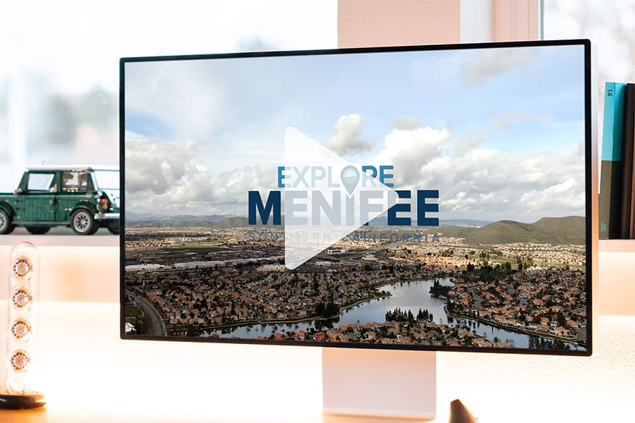 A computer monitor showcases a video thumbnail with Explore Menifee over a cityscape and mountains, capturing the spirit of tourism. A toy car and clear speakers are arranged neatly on the desk beside it.