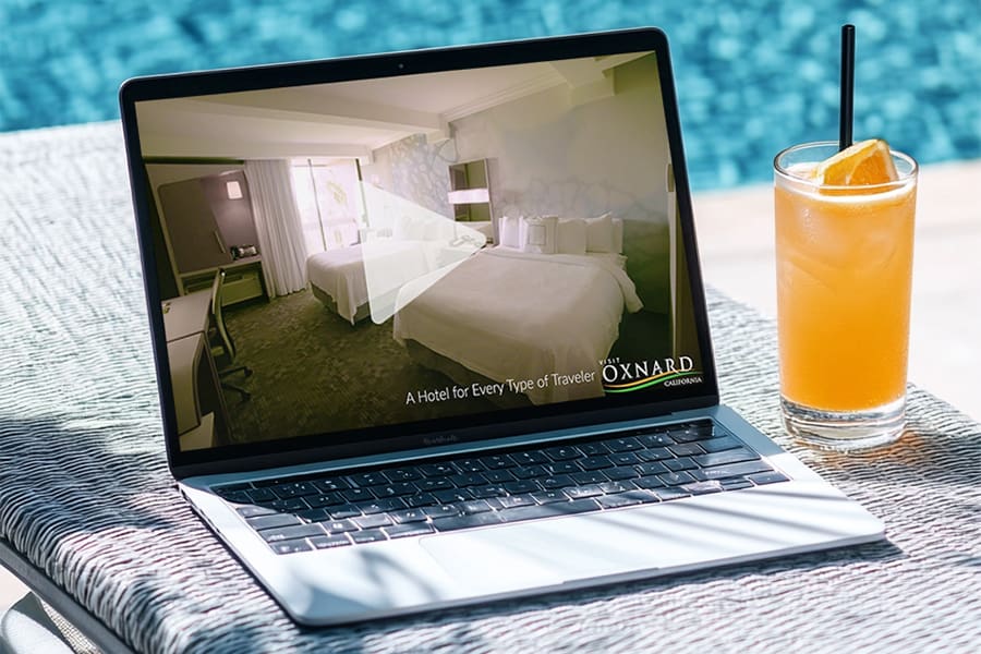 A laptop streaming a Hotels Video sits on a lounge chair beside a pool, capturing the essence of Oxnard Hotels. Nearby, a chilled glass of orange juice with a straw adds to the relaxed ambiance.