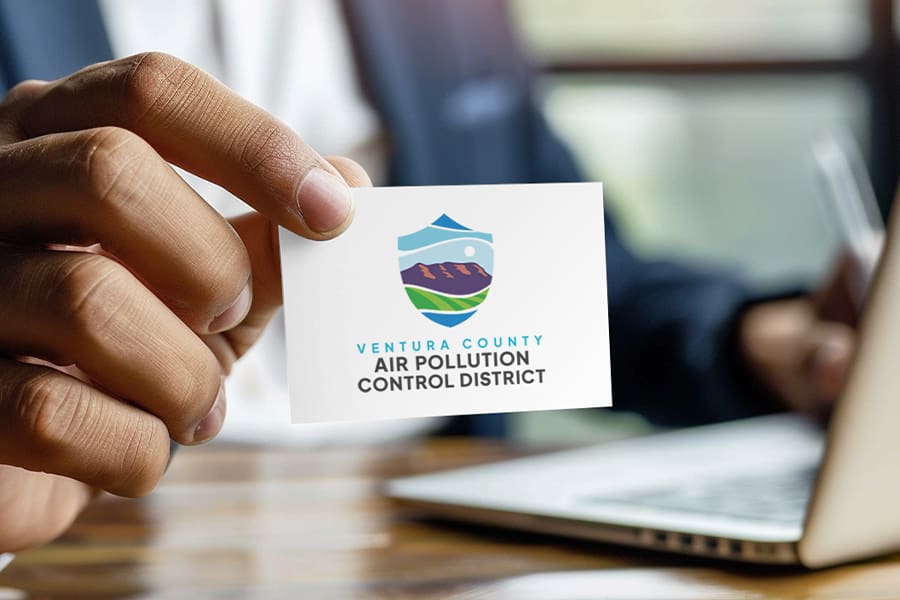 VCAPCD_Portfolio_02 Close-up of a person holding a business card displaying the Ventura County Air Pollution Control District logo, featuring stylized mountains and sun. The branding shines against the backdrop of a laptop and a softly blurred scene.