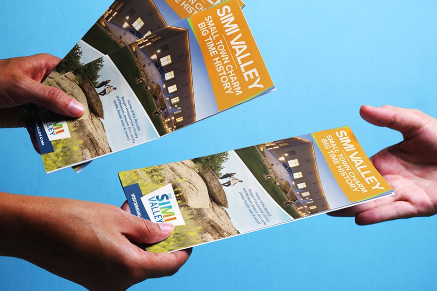 VisitSimiValley_RackCard_Portfolio_Mockup-2 Two hands exchanging a tourism brochure titled Simi Valley: Small Town Charm, Big Time History against a blue background.