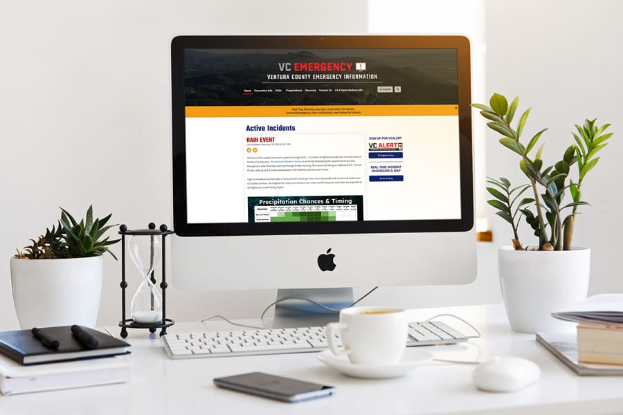 A computer on a desk showcases the VC Emergency Website. Surrounding it are three potted plants, an hourglass, a smartphone, and a steaming cup of coffee, creating a focused yet relaxed atmosphere.