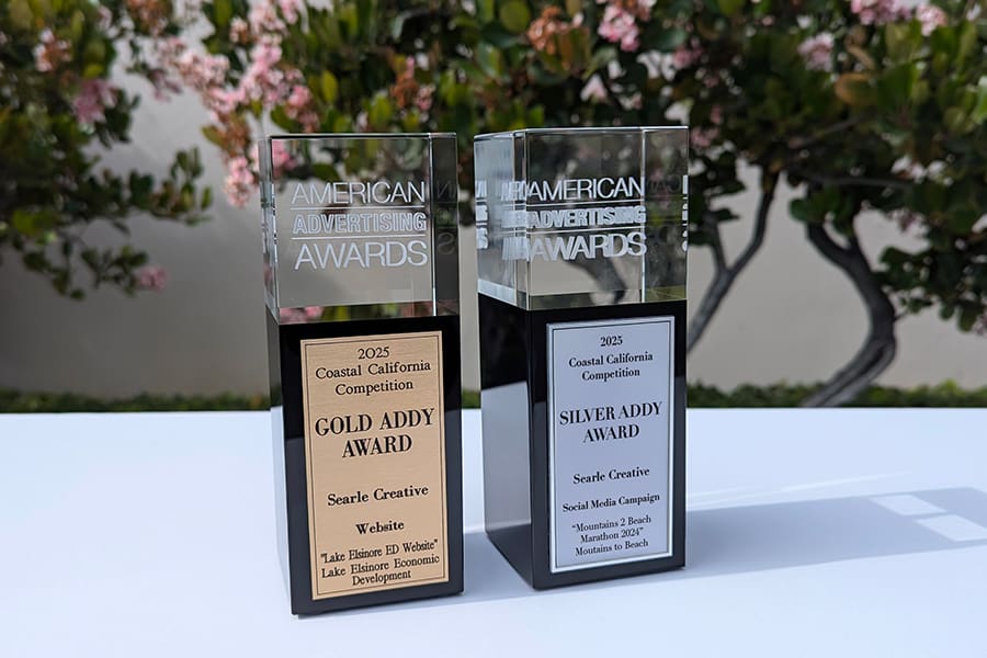 Addy2025_Gold-Silver_Award-Horizontal_Web Two rectangular glass trophies labeled American Advertising Awards (ADDYs), with gold and silver plaques, are displayed outdoors in front of greenery—celebrating Searles 2025 achievements.