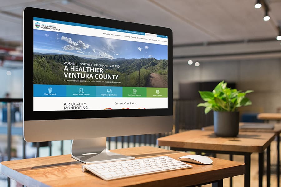 A computer monitor on a wooden desk displays the Ventura County Air Pollution Control District website in a modern, open office setting.